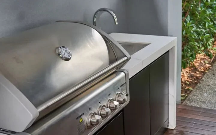 Stainless steel outdoor grill with temperature gauge and control knobs beside a sink