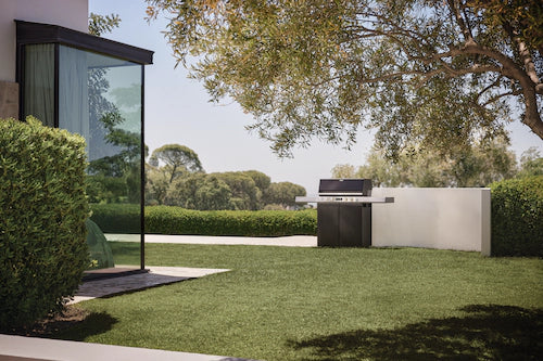 An outdoor kitchen setup featuring a black gas grill positioned next to a white wall, set in a landscaped backyard with a green lawn, hedges, and a modern glass-walled structure under a tree canopy.