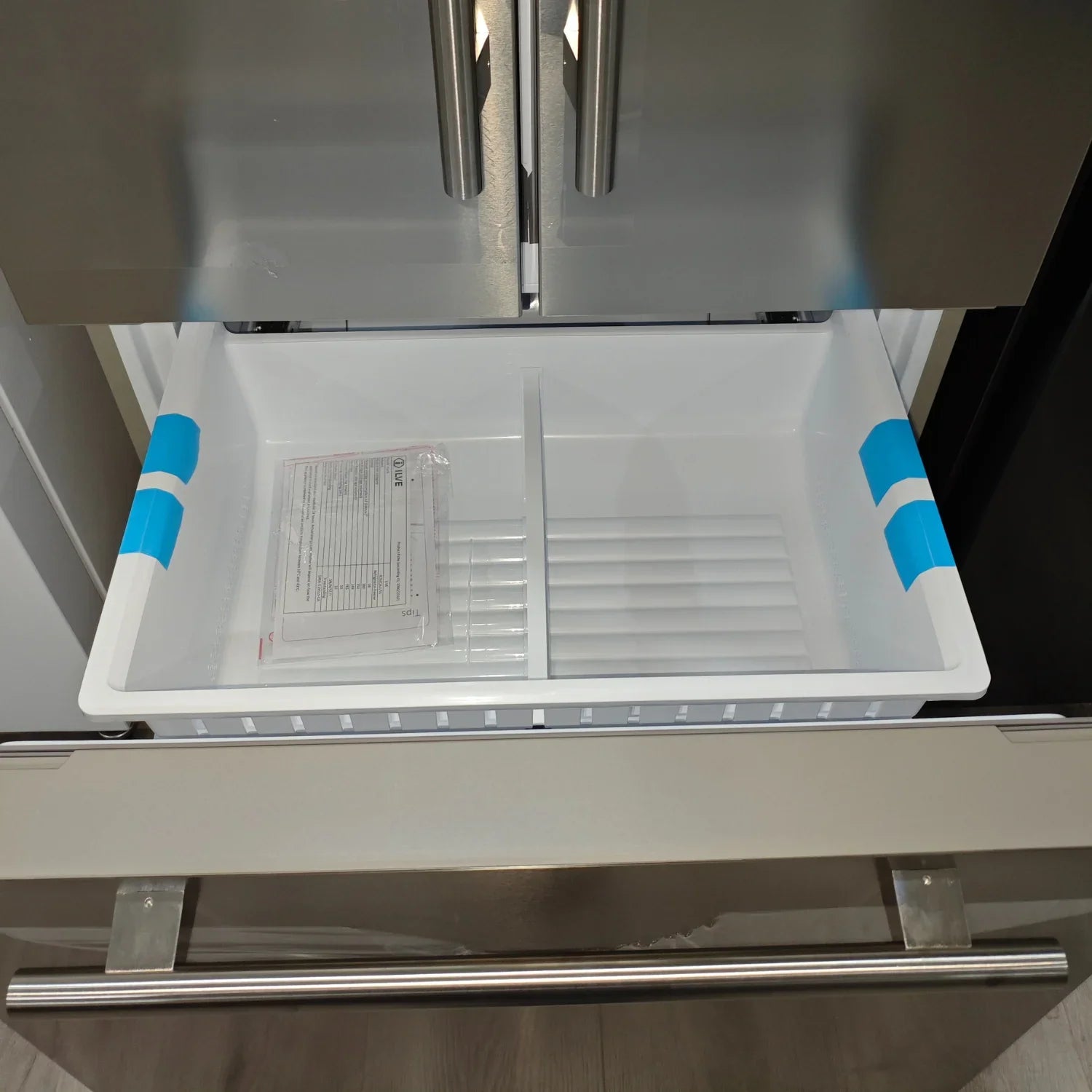 Stainless steel refrigerator with open drawer, white interior, blue tape, and instruction manual inside