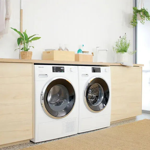 White front-load washer and dryer set in modern laundry room with wood cabinetry and plants