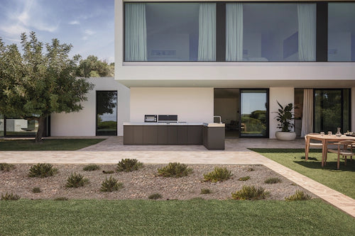 A modern outdoor kitchen island with dark gray countertops and a built-in sink, set in a landscaped backyard in front of a contemporary white house with large windows.