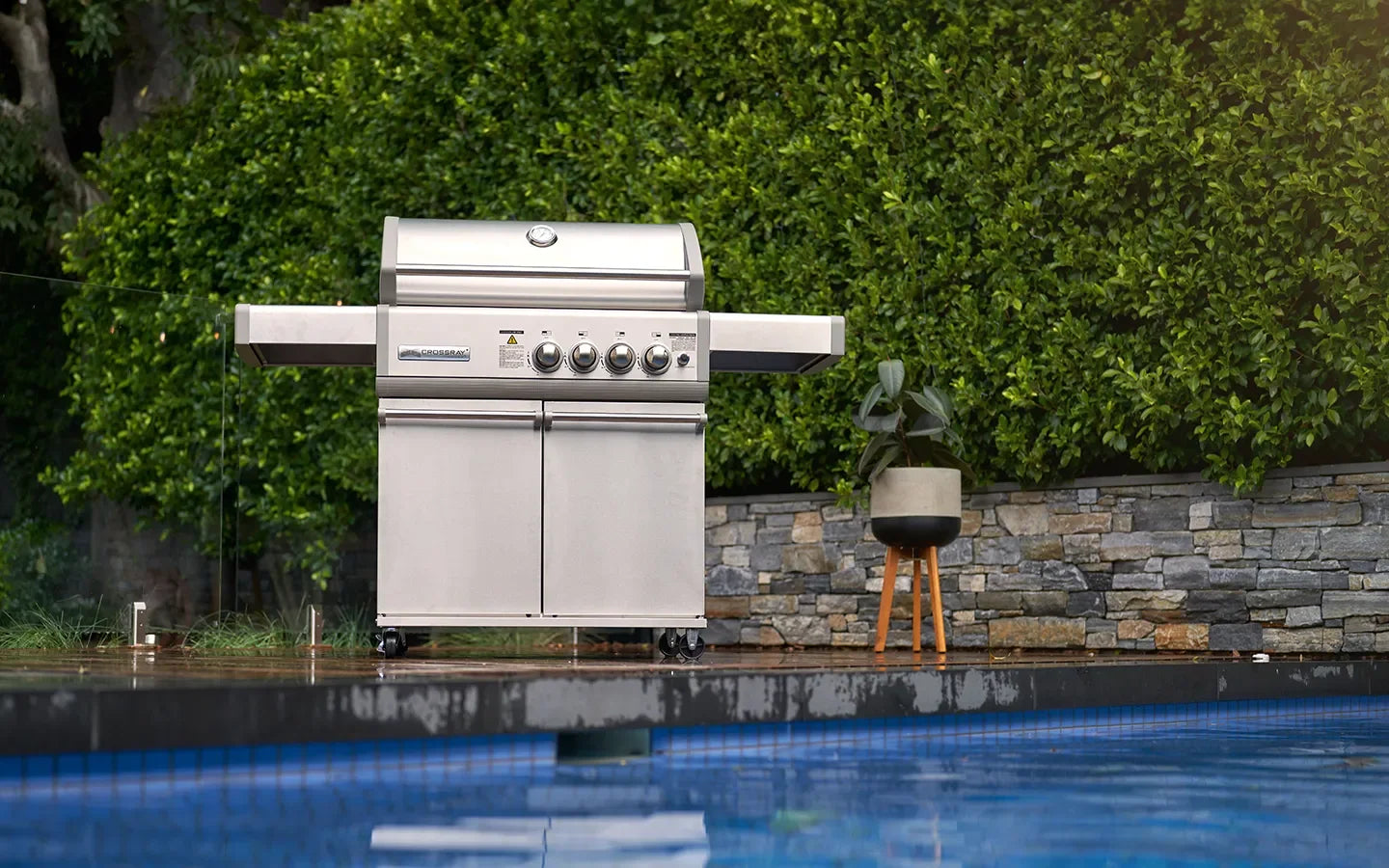Stainless steel outdoor grill by poolside with green hedge backdrop