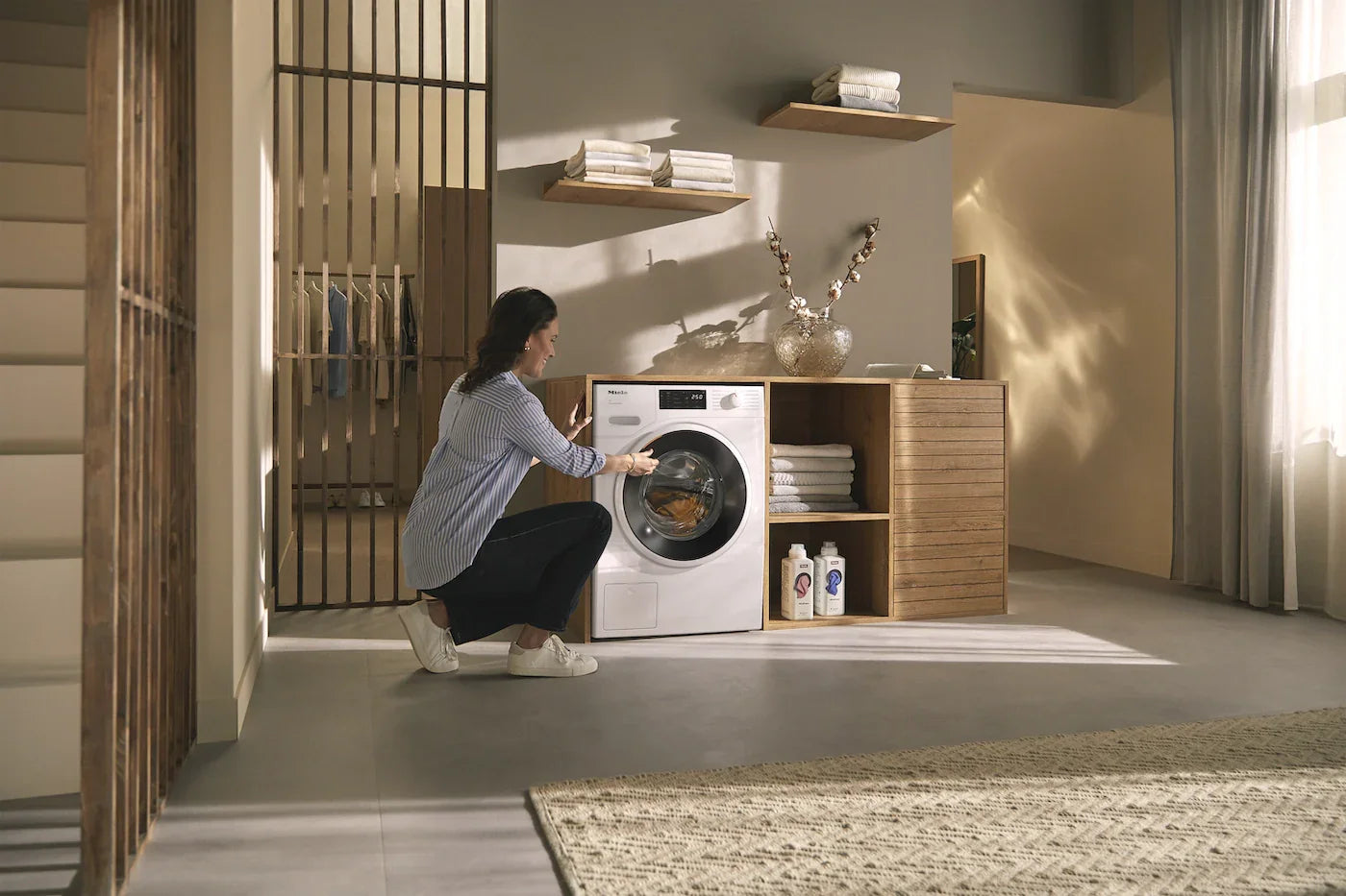 Woman loading white front-load washing machine in modern laundry room with wooden shelves and natural light
